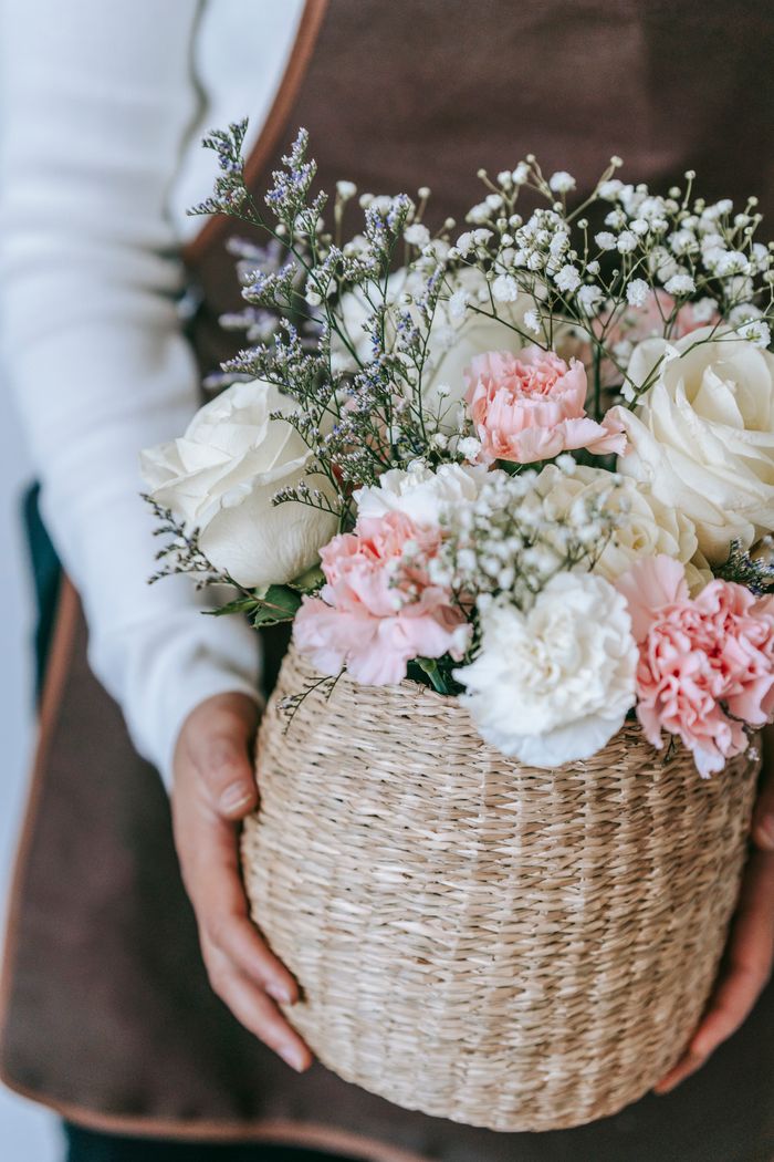 Crop anonymous florist in apron carrying wicker basket full of fresh gypsophila and pink and white roses
